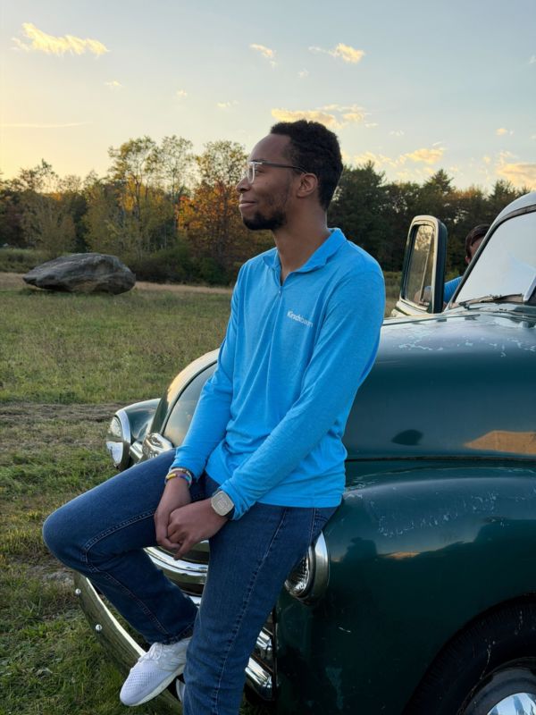 Malik sitting on the bumper of a vintage green Chevy truck in a field at sunset